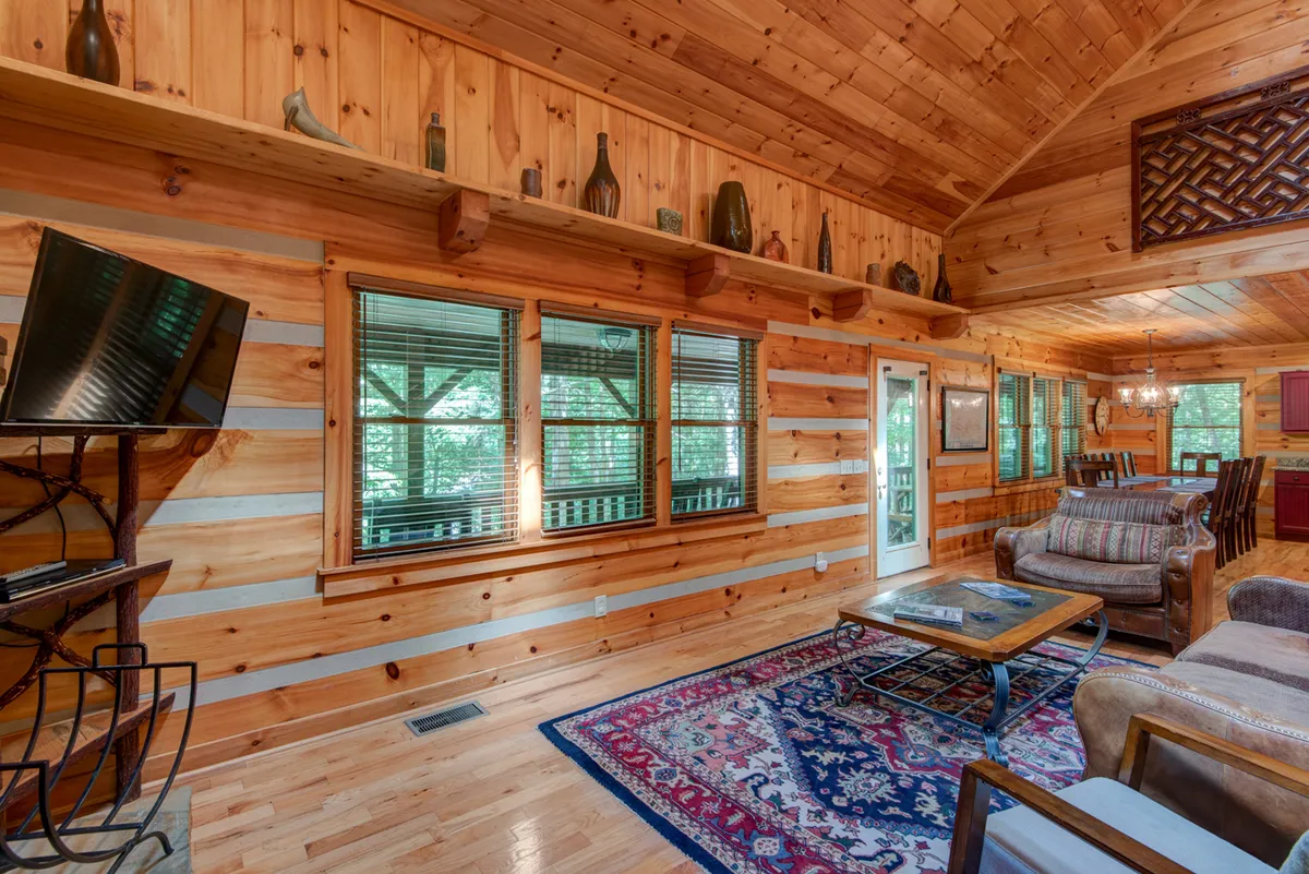 A view of the lodge’s main living area featuring a high wooden ceiling, expansive windows that flood the space with light, a cozy seating arrangement with a rug, and a shelf lined with bottles. The large windows offer glimpses of lush greenery outside, suggesting a strong connection to the surrounding landscape.