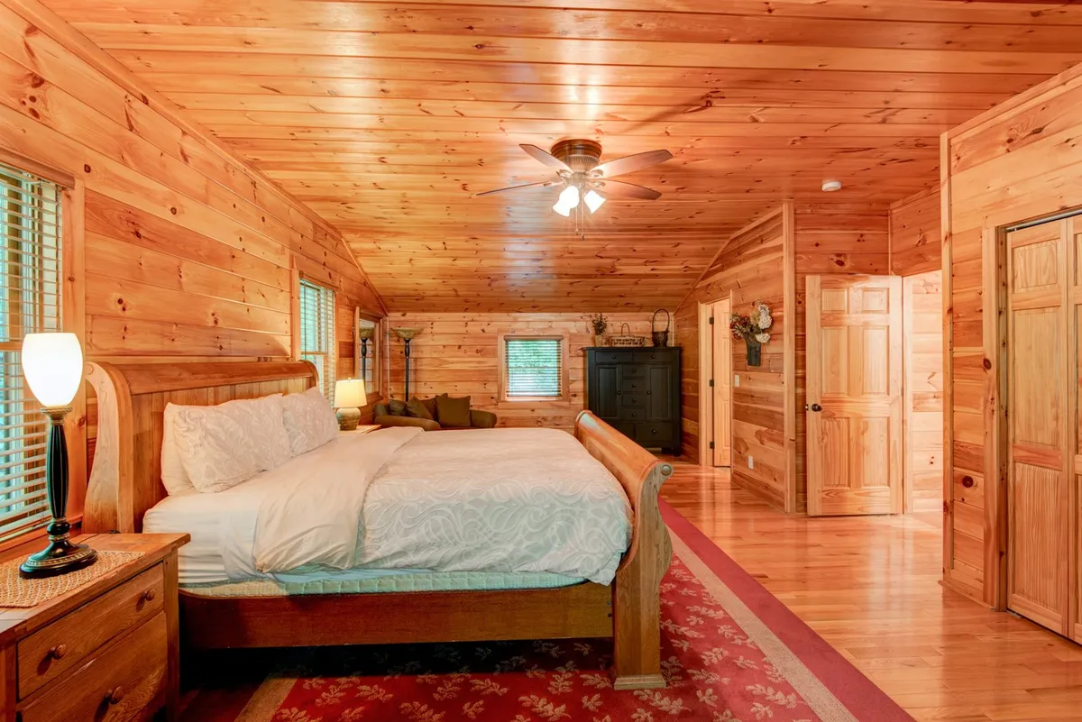 A cozy bedroom featuring a wooden bed, bedside lamp, plush rug, comfortable sofa, and wood-paneled walls. Natural light pours in through the windows, creating a calm and inviting space.