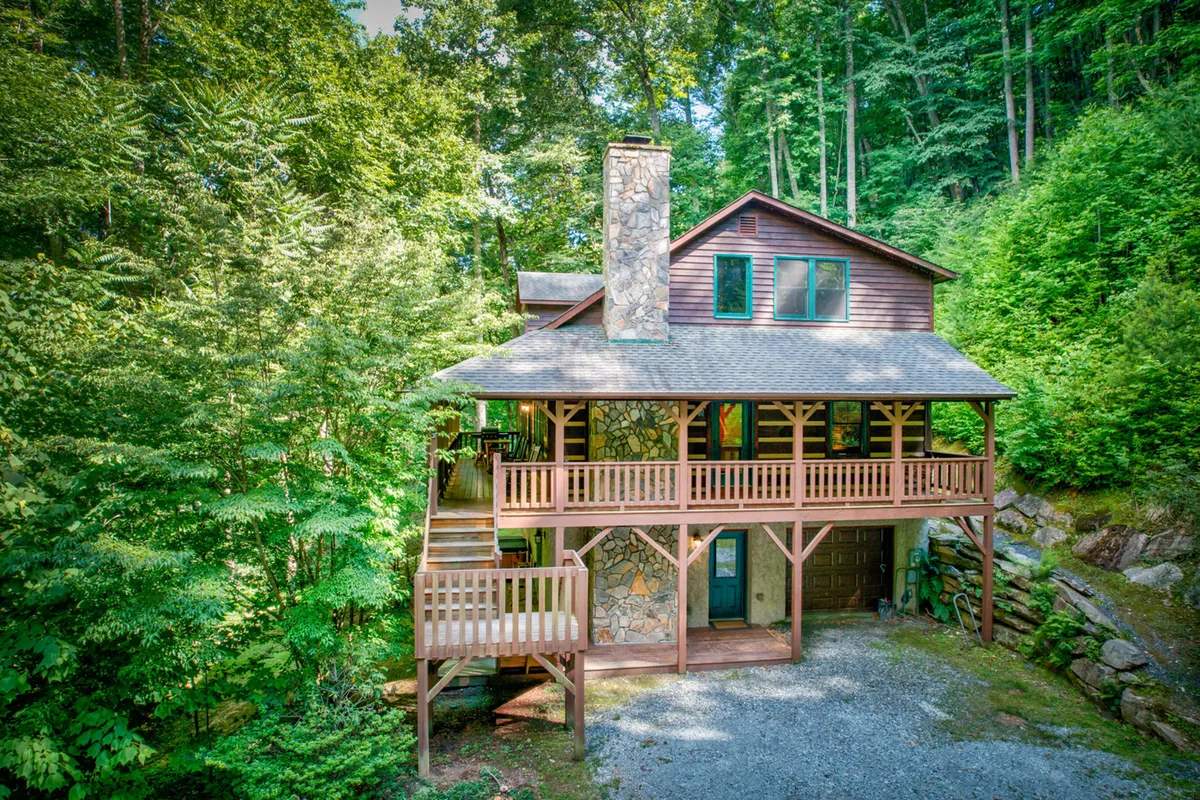 The image features the exterior of Little Creek Lodge, showcasing a classic wooden deck perfect for relaxation, a striking stone chimney hinting at cozy fires within, and lush greenery surrounding the property, inviting guests to enjoy the tranquility of nature. The front steps welcome visitors into the cabin, suggesting a warm and inviting retreat.
