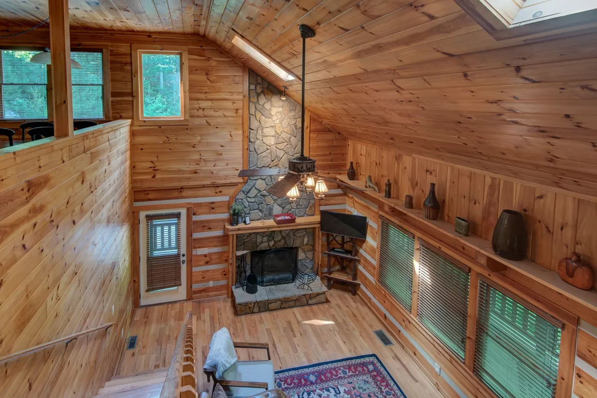 A warm living area featuring a rustic stone fireplace, wooden ceiling, and natural textures. Large windows offer a glimpse of the outdoors, filling the space with light and inviting nature in. Cozy seating is arranged for socializing or quiet respite after a day outdoors.