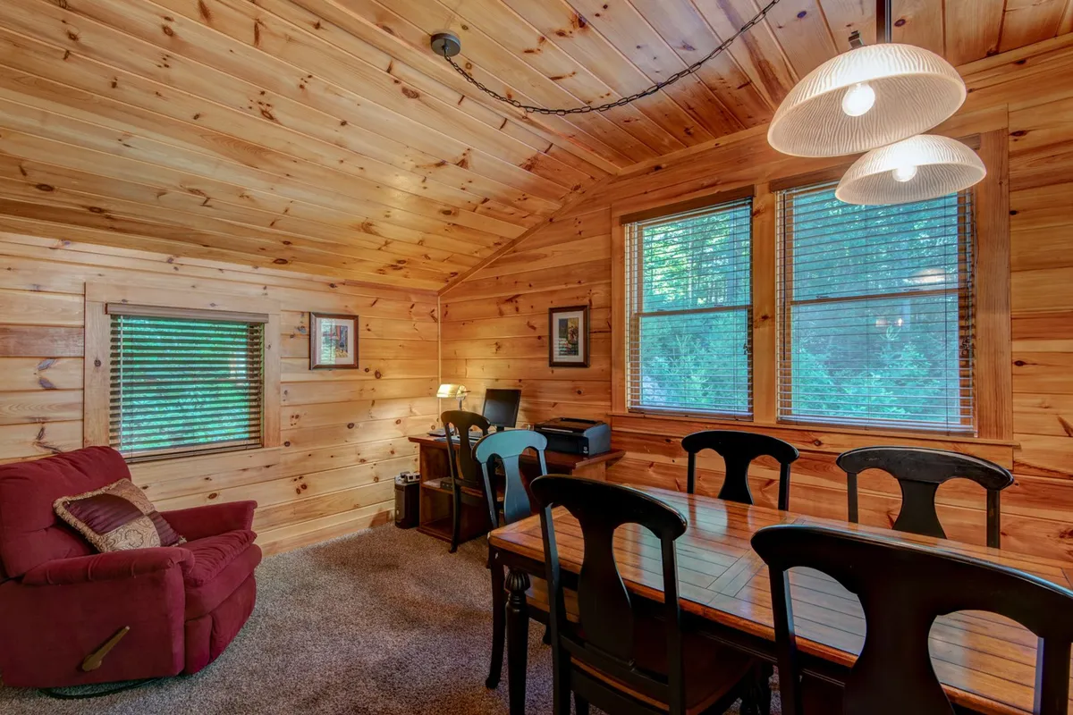 A warm, wood-paneled room with a sturdy wooden table, a plush red armchair, and large windows looking out to the greenery beyond. The space feels equally suited for shared meals or quiet reflection after outdoor excursions.