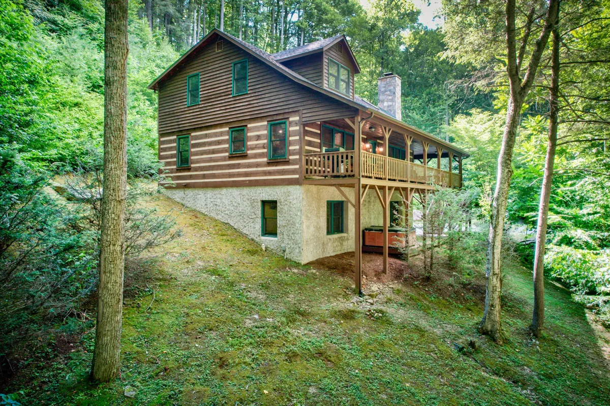 A wide wooden deck wraps around the house, surrounded by lush greenery and mature trees. The spacious deck is equipped with outdoor seating, inviting guests to relax and enjoy the sights and sounds of nature, while the house’s rustic wooden exterior blends seamlessly with the environment.
