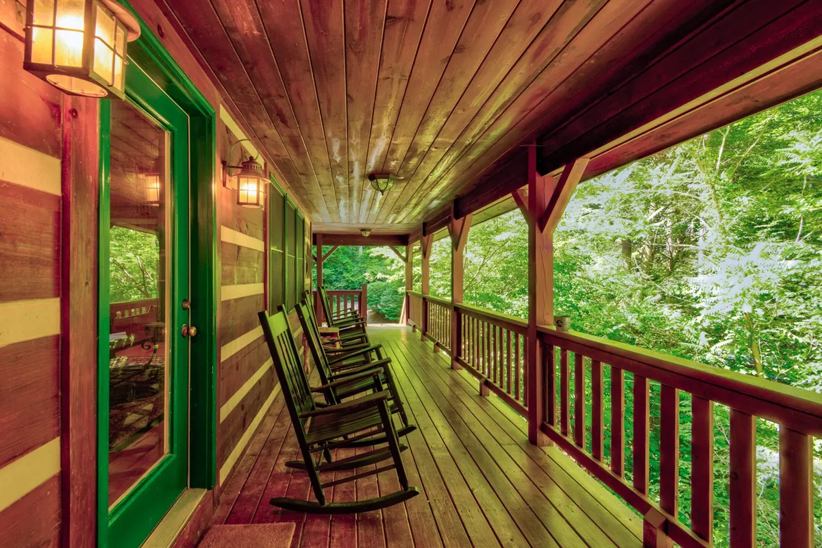 A covered porch with classic rocking chairs overlooks vibrant greenery. The welcoming green door and sturdy wooden railing add charm, while the tranquil surroundings invite relaxation and socializing with family or friends in the fresh mountain air.