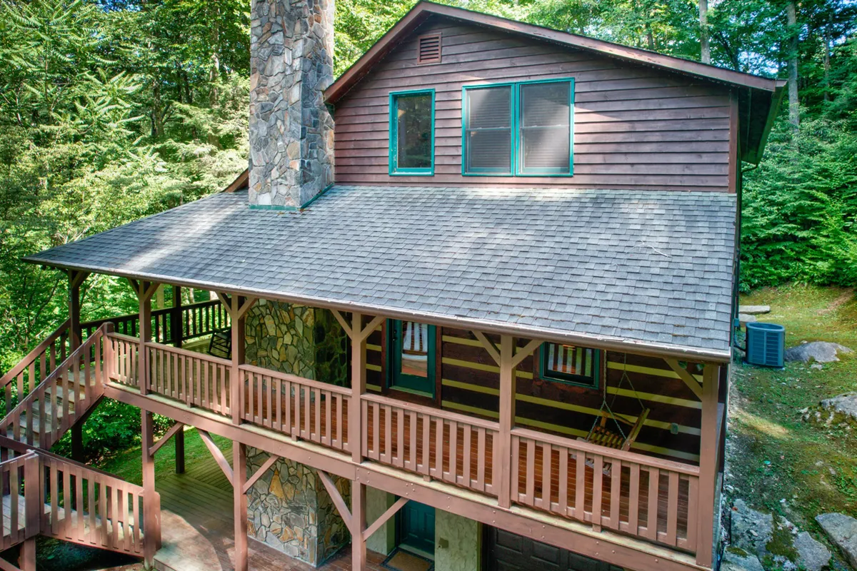 This image presents the exterior of Little Creek Lodge, highlighting a stone chimney, a wooden balcony, lush green trees, and expansive windows. The scene evokes a welcoming, cozy atmosphere ideal for group gatherings and relaxation in nature.