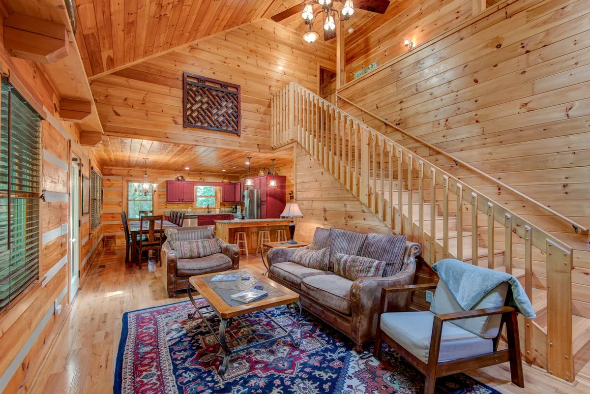 A welcoming living area with a beautiful wooden staircase, comfortable seating arranged around a coffee table, warm wood tones, and a dining table visible in the background. The space exudes rustic charm and comfort, perfect for relaxing together after exploring the outdoors.