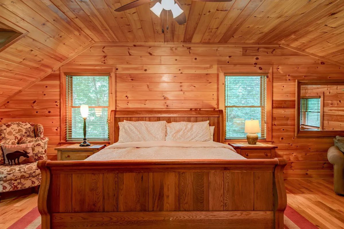 A serene bedroom featuring a wooden bed frame, cozy armchair, soft lighting from lamps, and windows that let in gentle natural light. The room's design emphasizes comfort and relaxation, with neutral tones and inviting textures.