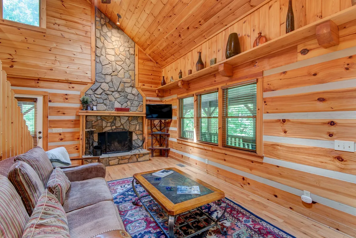 A warm and inviting den featuring a prominent stone fireplace, large windows flooding the space with natural light, wooden accents throughout, and a comfortable coffee table gathering area. The seating arrangement is perfect for groups to relax, watch television, play games, or share stories after a day of adventure.
