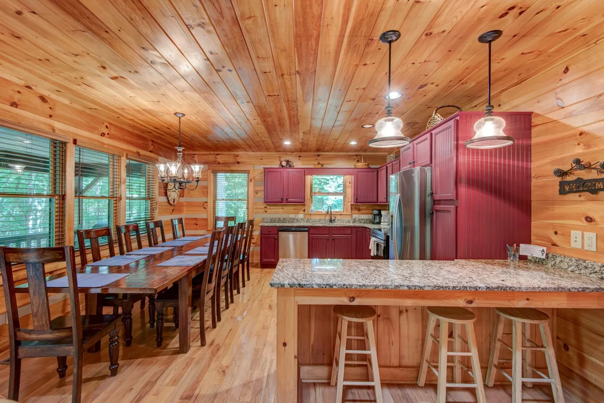 An inviting dining area features a long wooden table set for a group, surrounded by kitchen cabinetry and granite countertops. Hanging lights create a welcoming glow, and the open layout flows seamlessly from the kitchen to the dining space.
