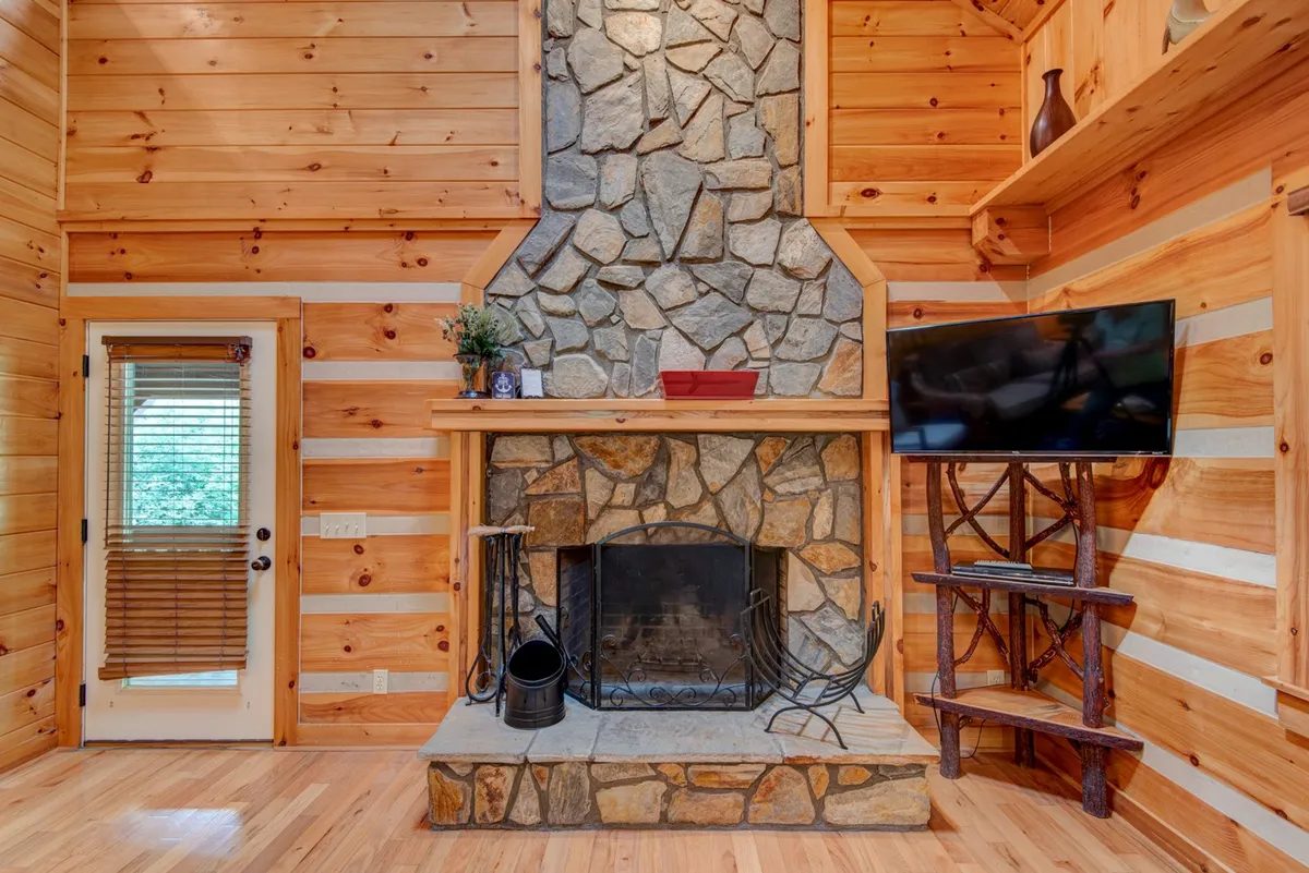 A welcoming family room accented by rustic wooden beams, a substantial stone fireplace, a large television above, and cozy seating. The decor is homey with a touch of greenery, inviting guests to unwind, watch movies, or enjoy intimate conversations together.