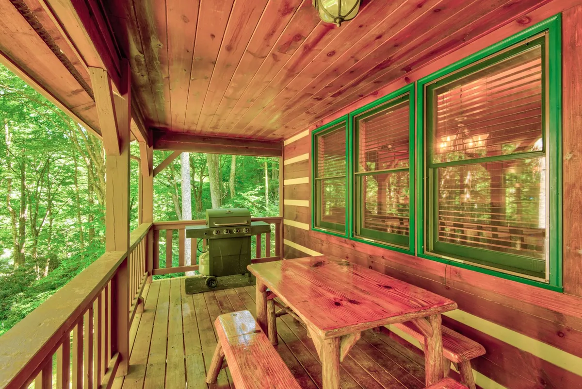 A large covered deck with a wooden table and a grill overlooks lush greenery. The setup is ideal for outdoor dining, with plenty of space for guests to gather and enjoy the mountain environment.