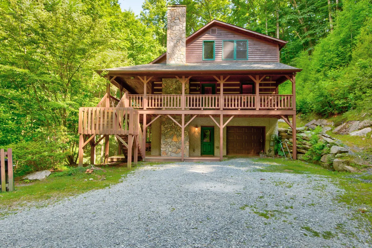 A rustic yet inviting view of the home's exterior, featuring a wooden balcony, a prominent stone chimney, and a cheerful green front door, all surrounded by lush greenery. The setting hints at opportunities for enjoying coffee on the porch and gathering with family outdoors.