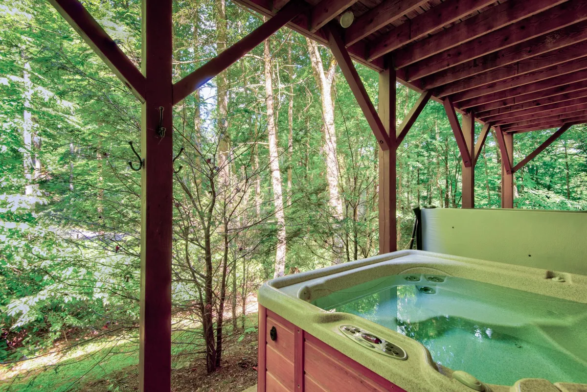 An exterior view of the lower deck featuring a bubbling hot tub surrounded by tranquil trees and a wooden structure. The setting is serene and private, ideal for soaking under the stars or warming up after a day outdoors.